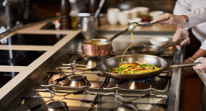 Person cooking vegetables in a pan on a stovetop with oil being added.
